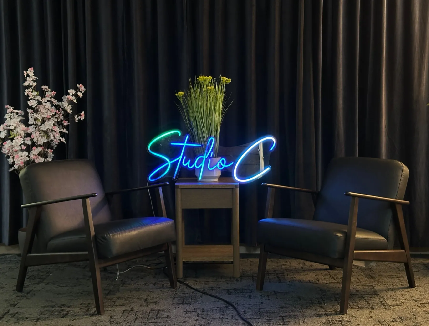 Two black armchairs in front of dark velvet curtains with a blue and green Studio C neon sign